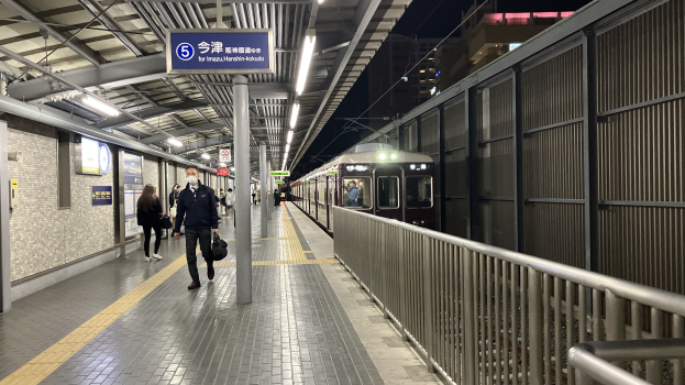 Ein Zug fährt auf einem Bahnsteig ein, mit Menschen, die gehen, einem Geländer auf der rechten Seite, Gebäuden im Hintergrund und Lampen mit einer Anzeigetafel oben auf dem Bahnsteig.