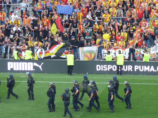 Eine Gruppe von Polizisten in Helmen und mit Waffen in der Hand läuft über ein Fußballfeld, während im Hintergrund Menschen mit Fahnen und Spruchbändern an Gittern stehen.
