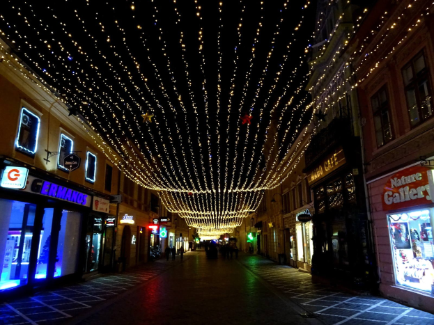 Eine festlich geschmückte Stadtstraße bei Nacht mit Weihnachtsbeleuchtung, flankiert von Gebäuden mit erleuchteten Fenstern und Schildern, mit wenigen Passanten auf dem Gehweg gegen einen dunklen Hintergrund.