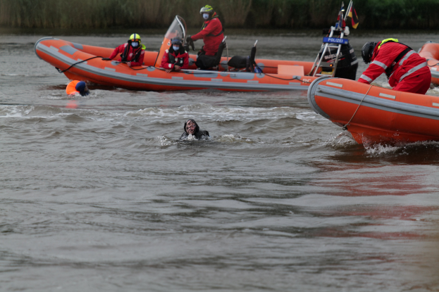 Gruppe von Menschen in einem aufblasbaren Boot auf einem Fluss, mit zwei Individuen im Wasser im Vordergrund und Vegetation im Hintergrund, alle tragen Schwimmwesten und Helme.