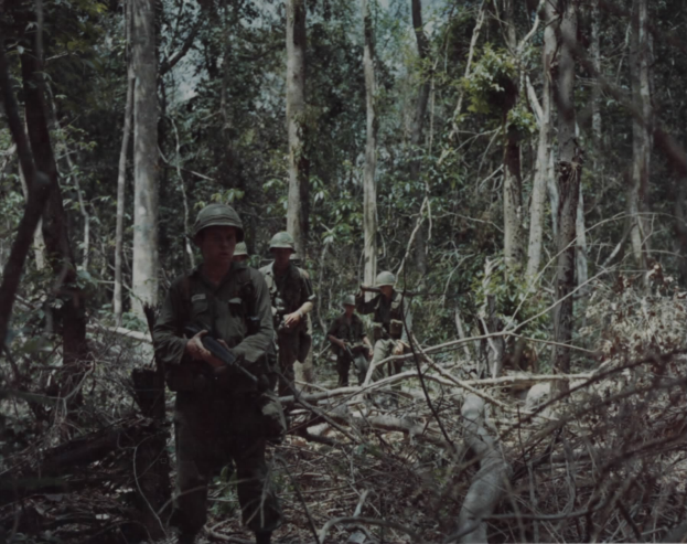 Eine Gruppe von Soldaten in Helmen und mit Gewehren durchquert einen grünen Wald mit hohen Bäumen und einem sichtbaren Himmel, mit Text am unteren Bildrand.