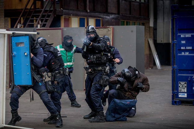 Eine Gruppe von Polizisten in Einsatzausrüstung steht vor einem Gebäude in Formation, wobei eine Person im Vordergrund liegt.