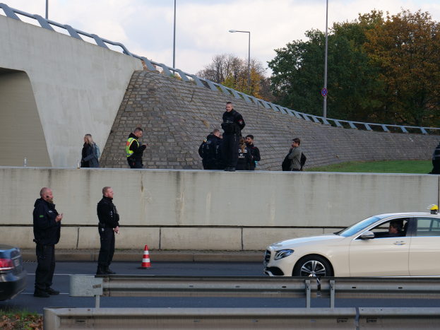 Eine Gruppe von Polizisten steht neben einem Auto auf der Straße, mit Verkehrskegeln, einer Begrenzung, Gras, einer Wand, Laternen, Bäumen und einem bewölkten Himmel im Hintergrund.