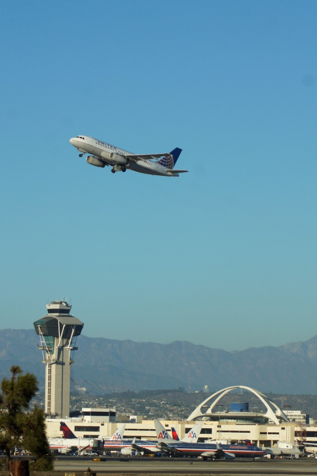 Eine Flughafenansicht mit einem weißen Kontrollturm und parkenden Flugzeugen im Vordergrund, Gebäuden im Hintergrund und einem Flugzeug am Himmel