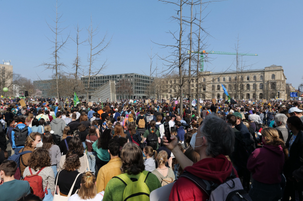 Eine große Menge Menschen bei einer Klimawandel-Demonstration vor einem Gebäude mit Bäumen und einem klaren Himmel im Hintergrund.