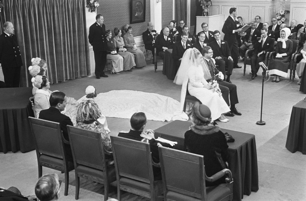Schwarzes und weißes Foto einer Hochzeitszeremonie mit sitzenden und stehenden Menschen, einem Mann mit Mikrofon, einer Frau mit einem Blumenstrauß, einem Podium mit Mikrofon, Blumengestecken, einem Vorhang, einem gerahmten Foto an der Wand und einer Tür im Hintergrund.