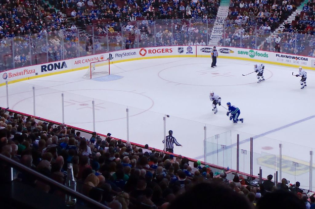 Vier Männer spielen Hockey auf einem Eisrink, mit zwei in der Nähe des Tors und des Zauns stehend, Zuschauern auf Stühlen im Vorder- und Hintergrund und einer Treppe im Hintergrund.