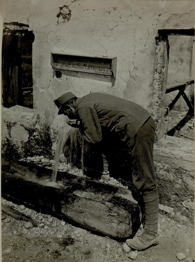 Ein Mann mit Mütze und Schuhen trinkt Wasser aus einem Brunnen vor einer Wand mit einem Fenster und einem Schild.