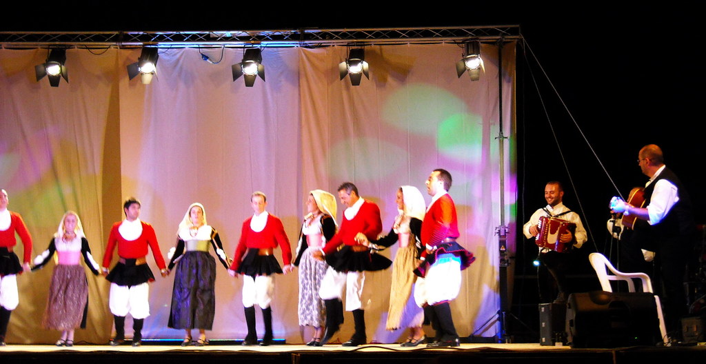 Performer auf der B├╝hne mit einem Vorhanghintergrund, Scheinwerfer ├╝berkopf, Musiker, die Instrumente spielen, und ein weißer Stuhl in der rechten unteren Ecke.