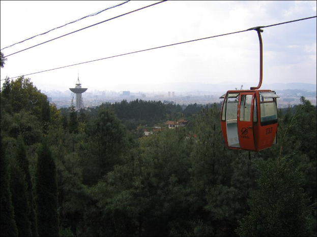 Eine Seilbahn bewegt sich vorne mit einer Stadtlandschaft, einschließlich Gebäuden, einem Turm und Bäumen, unter einem bewölkten Himmel im Hintergrund.