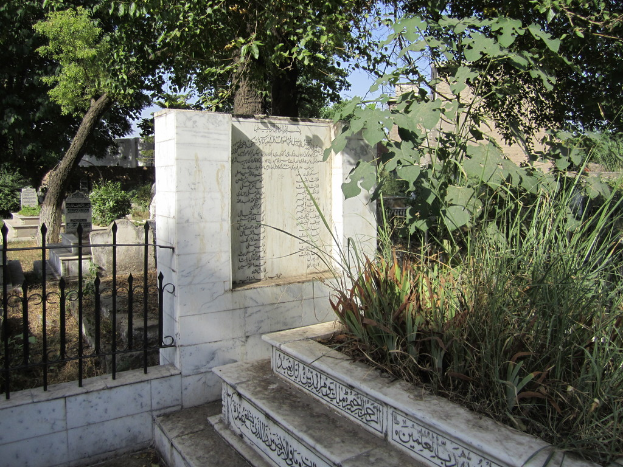 Ein Gedenkschild neben einem Geländer in einem Friedhof, mit Treppen unten, Gras und Bäumen auf der rechten Seite, Gräbern hinter dem Geländer und Gebäuden und Bäumen im Hintergrund.