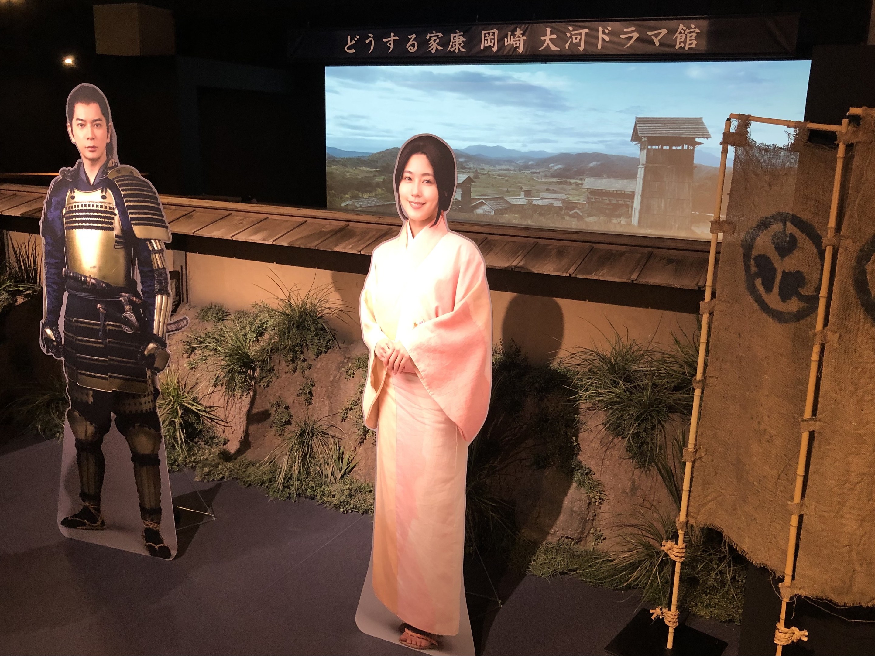 Zwei Menschen in traditioneller japanischer Kleidung - eine Frau in einem pinken Kimono und ein Mann in Samuraikleidung - lächeln gemeinsam in einer Inszenierung mit Pflanzen, einem Bildschirm und Beleuchtung, die auf eine Filmszene hinweist.