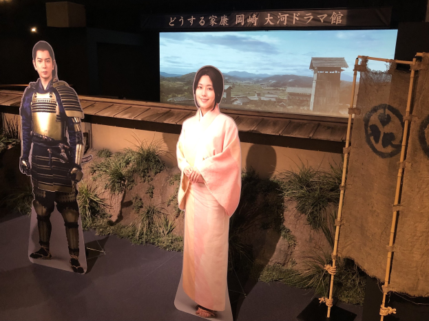 Zwei Menschen in traditioneller japanischer Kleidung - eine Frau in einem pinken Kimono und ein Mann in Samuraikleidung - lächeln gemeinsam in einer Inszenierung mit Pflanzen, einem Bildschirm und Beleuchtung, die auf eine Filmszene hinweist.