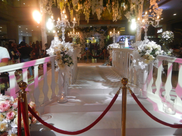 Eine Hochzeitszeremonie in einem Einkaufszentrum mit weißem Teppich, Geländern, Blumenschmuck, Kronleuchtern und Deckenbeleuchtung, bei der viele Menschen anwesend sind, einige mit Taschen.