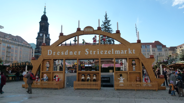 Ein belebter Weihnachtsmarkt in Dresden, Deutschland mit Menschen, die herumlaufen, einige mit Taschen, unter einem Torbogen, mit Bäumen und Gebäuden im Hintergrund unter einem bewölkten Himmel.