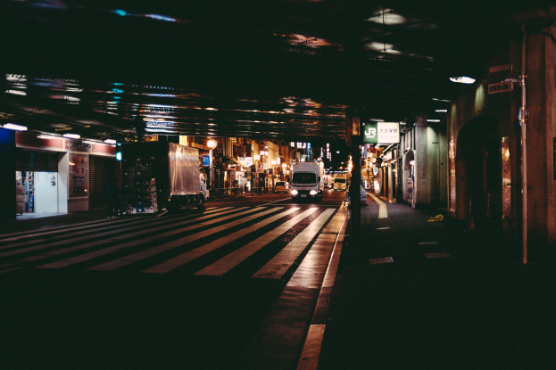 Eine Stadtstraße bei Nacht mit Fahrzeugen auf der Straße, Gebäuden auf beiden Seiten, beleuchteter Gegend und einer Brücke im Hintergrund.