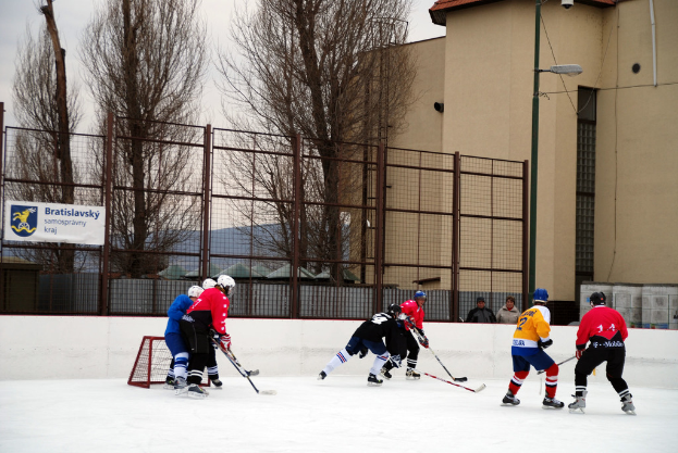 Personen spielen Eishockey auf einer Eisfläche mit Gebäuden, Bäumen, einer Straßenlaterne, einem Namensschild und Zäunen im Hintergrund unter einem Himmel.
