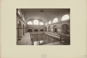 Schwarzes und weißes Foto eines Hallenbads mit Geländern, Bänken, gewölbten Wänden und Beleuchtung, das Text am unteren Rand zeigt.