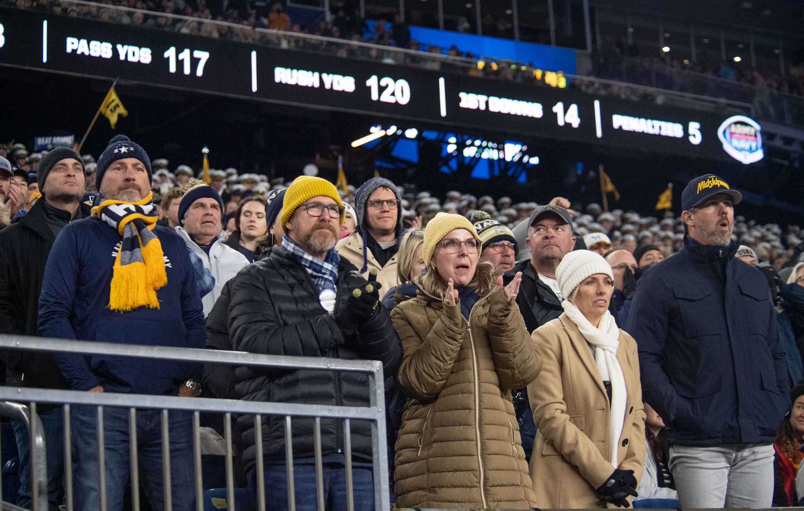 People standing in front of a crowd at a football game, some wearing caps and scarves, with railings in the foreground and illuminated boards displaying text and numbers in the background.