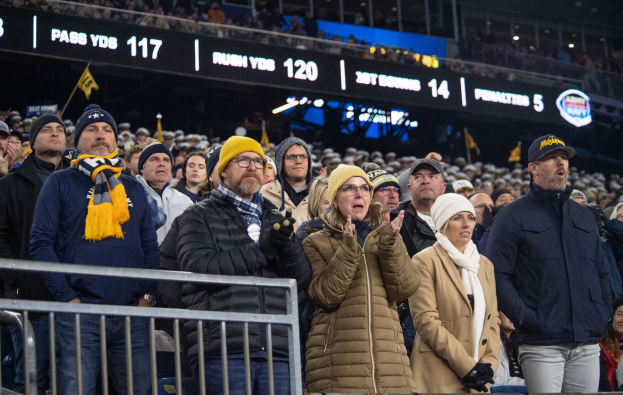 People standing in front of a crowd at a football game, some wearing caps and scarves, with railings in the foreground and illuminated boards displaying text and numbers in the background.