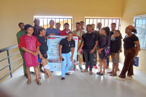 Eine Gruppe von Menschen, die zusammen in einem Raum stehen und ein Schild in der Mitte halten, mit einem Geländer auf der linken Seite, Fenstern und einer Wand im Hintergrund.