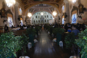 Eine Hochzeitszeremonie in einer Kirche mit Sitzplätzen und Stehplätzen, Topfpflanzen im Gang, Skulpturen an den Wänden, Vorhänge an den Fenstern, Kronleuchter und ein Bildschirm im Hintergrund.