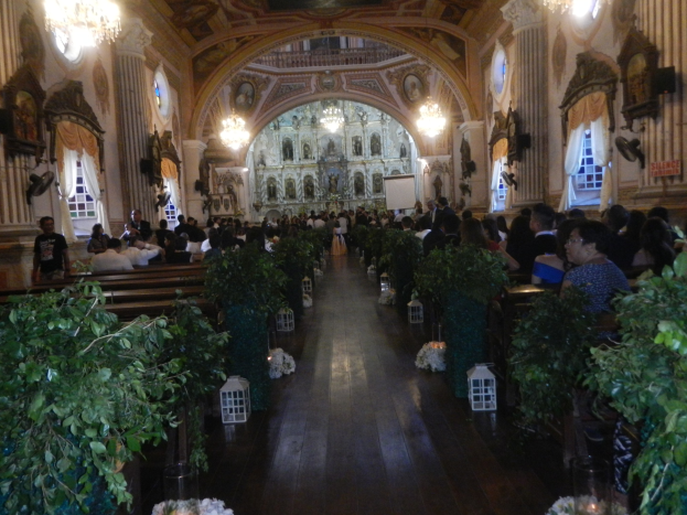 Eine Hochzeitszeremonie in einer Kirche mit Sitzplätzen und Stehplätzen, Topfpflanzen im Gang, Skulpturen an den Wänden, Vorhänge an den Fenstern, Kronleuchter und ein Bildschirm im Hintergrund.
