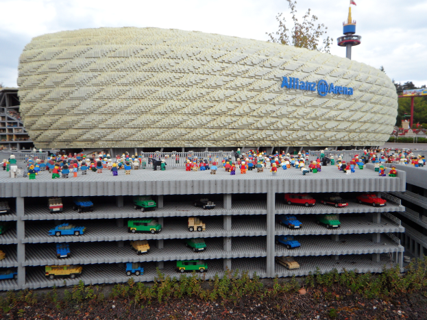 Eine große Lego-Struktur, die dem Allianz Stadion in München, Deutschland, ähnelt, mit einer Gruppe von Menschen davor, umgeben von Bäumen, Pfählen, einem Turm und Grünflächen, unter einem bewölkten Himmel.