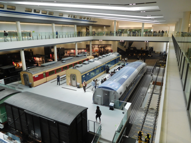 Großer Museumsraum mit verschiedenen Zügen in unterschiedlichen Größen und Farben, mit Menschen, die beobachten, und Geländern, Säulen und Deckenlampen.