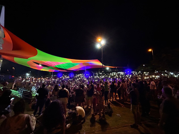 Eine große Menschenmenge steht vor einem hell erleuchteten Zelt in der Nacht, mit bunten Lichtern und Pfählen, die die Szene beleuchten, einem Gebäude auf der linken Seite und dem Nachthimmel im Hintergrund während des 2018er Kunstfestivals.