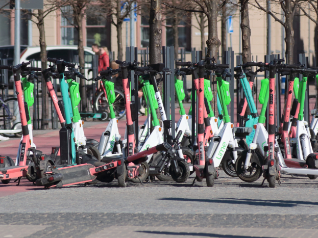 Eine Reihe von Elektro-Rollern, die an der Straße geparkt sind, mit einer Person im Vordergrund und Baumwipfeln und Gebäuden im Hintergrund.