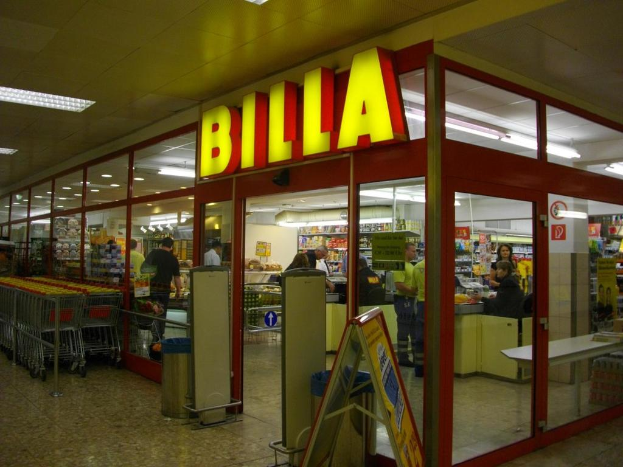 Ein Markt mit einem Eingang in der Mitte, Mülltonnen daneben, einer Reihe von Einkaufswagen auf der linken Seite und einigen Menschen drinnen, die unter Deckenbeleuchtung Produkte durchstöbern.