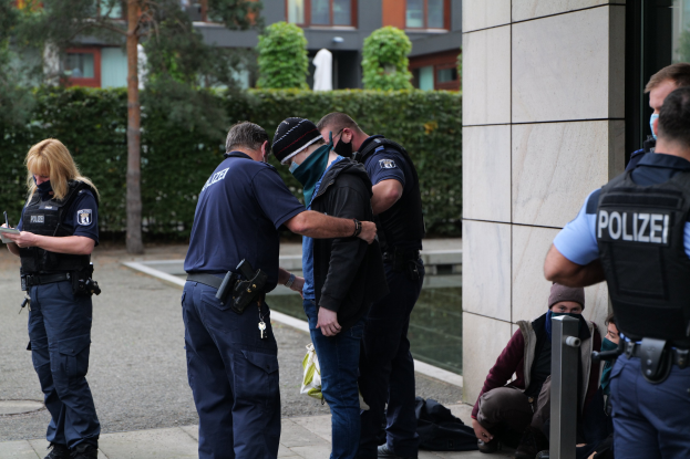 Deutsche Polizeibeamte stehen in einer Gruppe vor einem Gebäude, mit einer Frau, die ein Buch und einen Stift hält, und Menschen in der Nähe, vor einem Hintergrund aus Grünflächen und städtischen Strukturen.