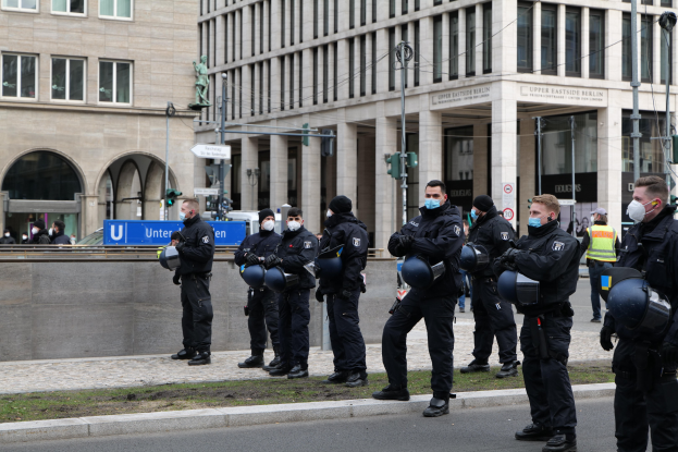 Eine Gruppe von Polizisten in schwarzen Uniformen und Masken steht vor einem Berliner Gebäude mit Glasfenstern und Säulen, einige halten Helme, mit Laternenmasten, Verkehrsampeln, Texttafeln, einer Statue und anderen Menschen in der Szene.