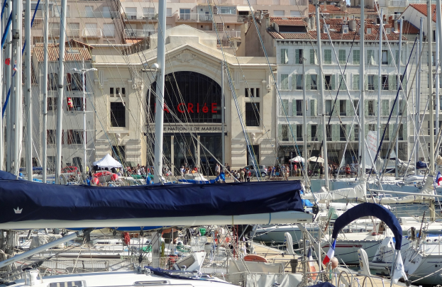 Marina mit zahlreichen Liegeplätzen, Gebäuden mit Fenstern und einem Namensschild im Hintergrund, Zelten und Menschen.