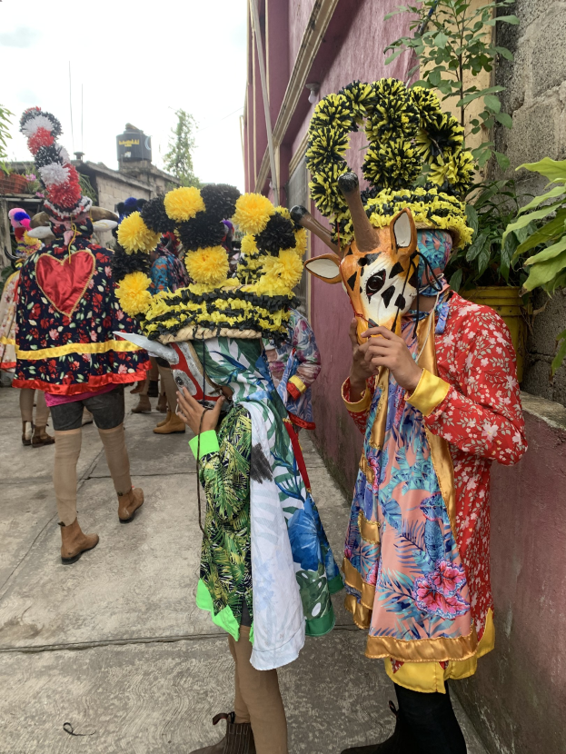 Eine Gruppe von Menschen in farbenfrohen Kostümen, die gemeinsam vor einem Gebäude stehen und verschiedene Gegenstände halten, mit Bäumen, Gebäuden und einem klaren blauen Himmel im Hintergrund.
