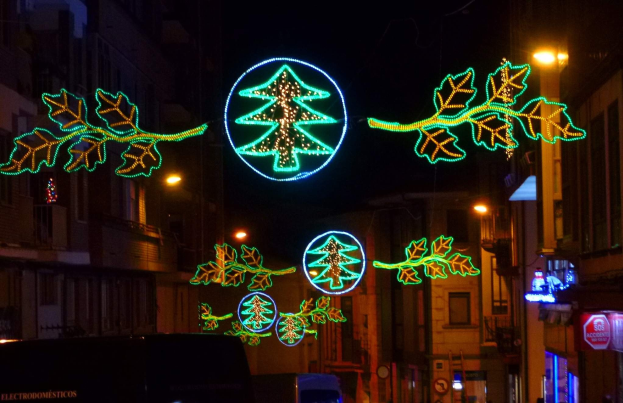 Eine städtische Straße, die mit festlichen Weihnachtslichtern geschmückt ist, die Blätter und andere dekorative Elemente zeigen, mit Fahrzeugen, die fahren und Gebäuden auf beiden Seiten.