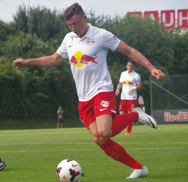 Ein professioneller Fußballspieler in einem roten Trikot kickt einen Ball auf einem grünen Feld mit Bäumen und einem klaren blauen Himmel im Hintergrund, wobei ein 'RB Leipzig'-Schild sichtbar ist.