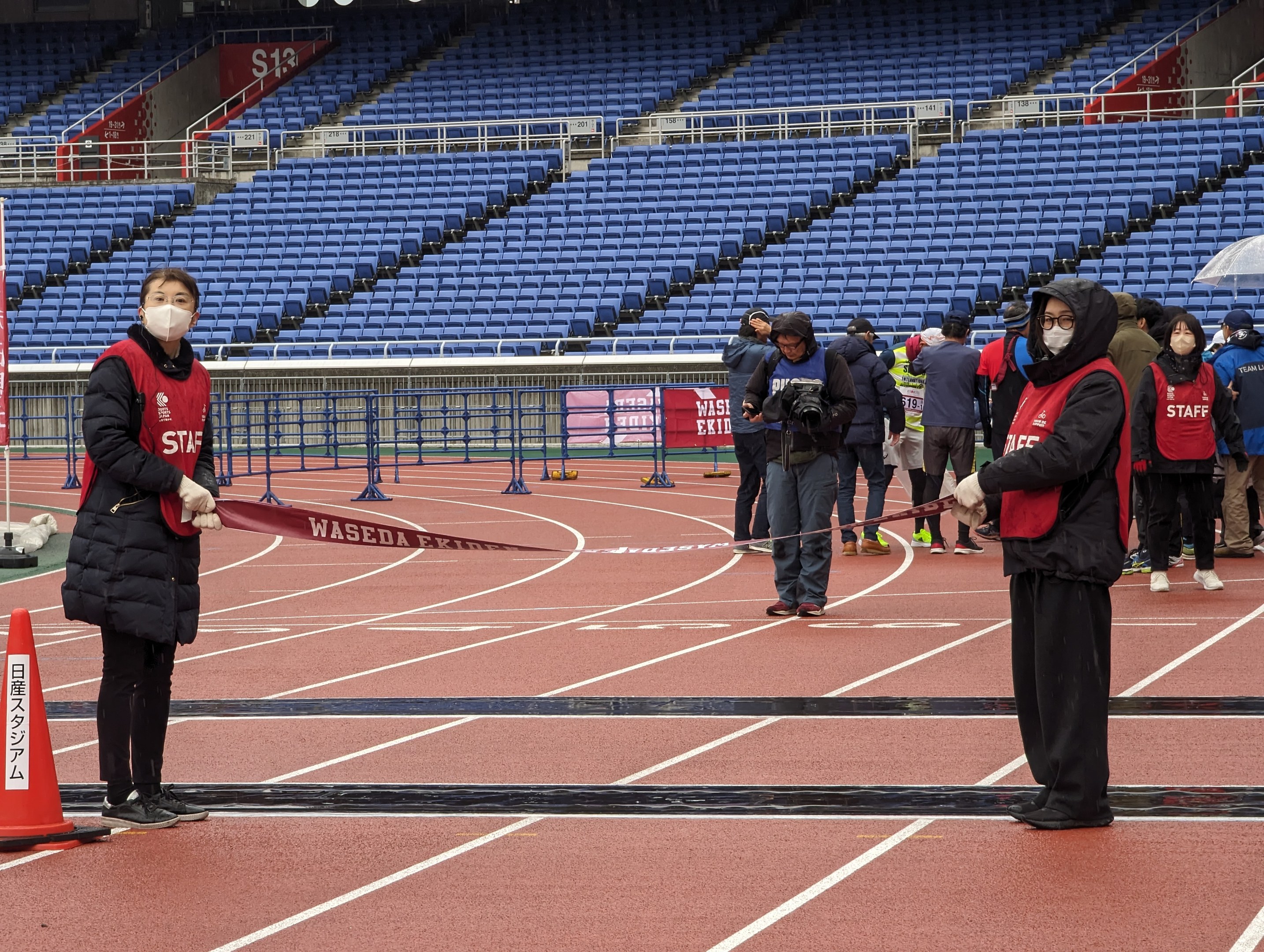 Gruppe von Menschen, die ein Band bei einem Rennen auf einer Bahn halten, mit einem Verkehrskegel links und Event-Aufbau im Hintergrund.