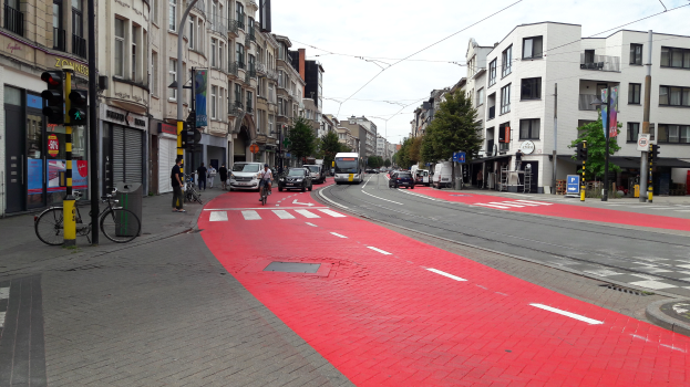 Eine Stadtstraße mit einem roten Fahrradweg, Fahrzeuge auf der Straße, Fußgönger auf dem Gehweg, Gebäude mit Fenstern auf beiden Seiten, Bäume und ein klarer blauer Himmel im Hintergrund.