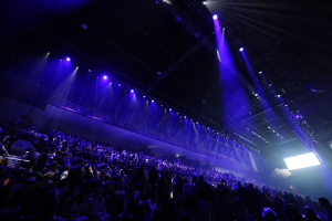 Eine große Menschenmenge sitzt vor einer Bühne mit Deckenbeleuchtung, mit einem Bildschirm auf der rechten Seite in einer Konzerthalle.