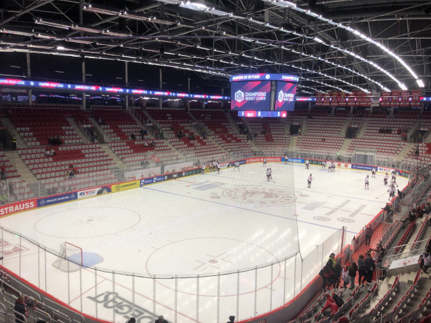 Eishockeyspiel in einer großen Arena zwischen den St. Louis Blues und Detroit Red Wings, mit Zuschauern auf Sitzen und Stehplätzen in der Nähe des Spielfelds umgeben von einem Zaun und Metallrahmen, unter einem Dach mit Deckenleuchten und einem Display.