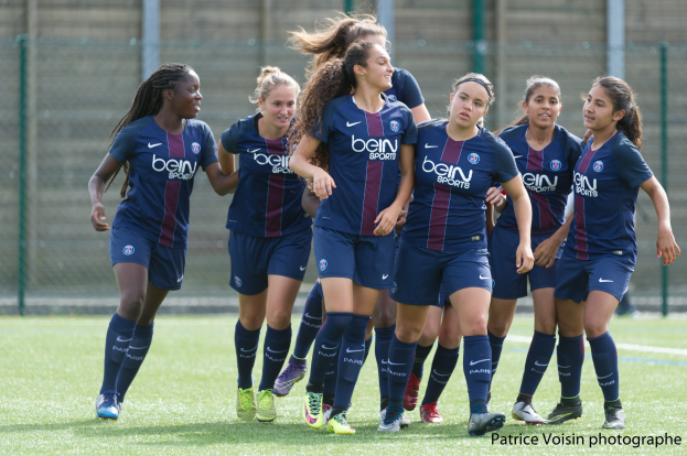 Eine Gruppe junger Frauen, die auf einem Rasenfeld mit Maschendrahtzaun und einer Wand im Hintergrund Fußball spielen, mit dem Text "Paris Saint-Germain Frauenfußball" in der rechten unteren Ecke.