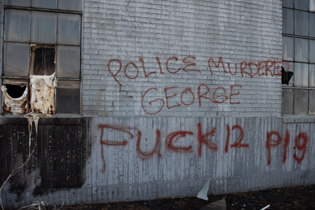 Ein Gebäude mit Graffiti, das "Polizei Mord George Fuck 12 P19" auf der Seite trägt und Fenster sowie verschiedene Gegenstände vor ihm zeigt.