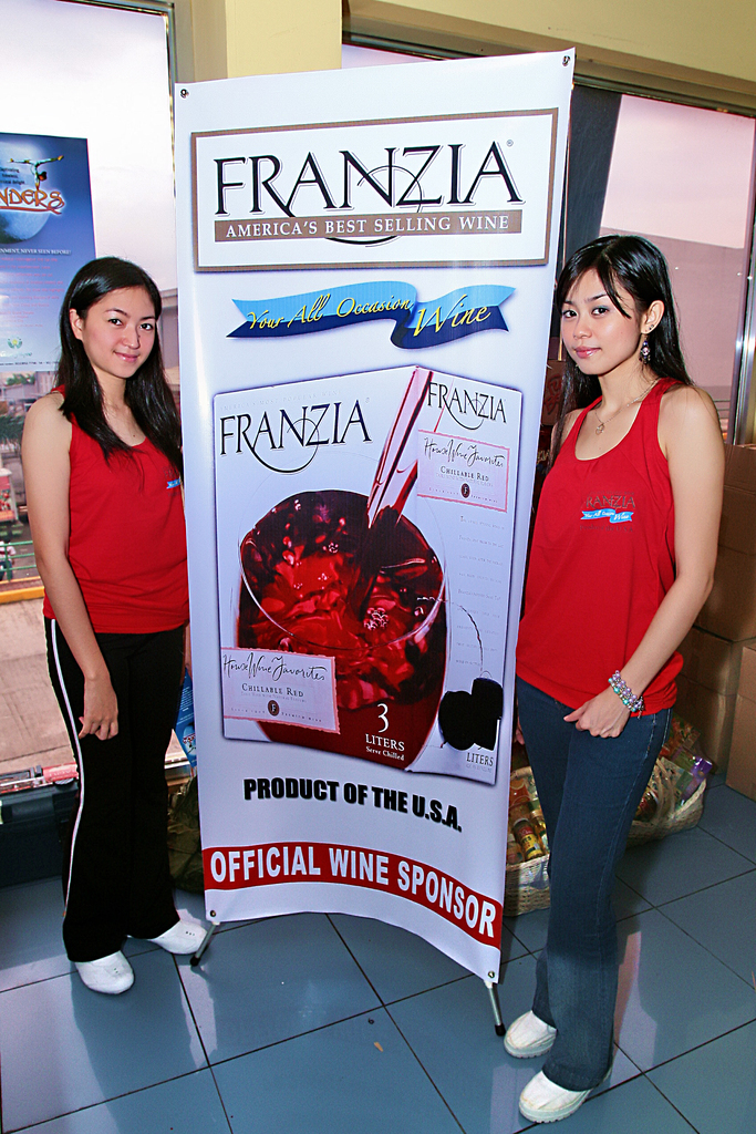 Zwei Frauen neben einem Plakat f├╝r Franzia, Amerikas bestverkaufenden Wein, mit einem Glasfenster im Hintergrund.