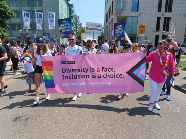 Eine bunte Gruppe von Menschen marschiert die Straße entlang, hält ein Banner mit der Aufschrift 'Vielfalt ist eine Tatsache, Inklusion ist eine Wahl', wobei einige Mützen und Schutzbrillen tragen und andere zusätzliche Banner halten, vor dem Hintergrund von Gebäuden, Bäumen und einem klaren blauen Himmel.