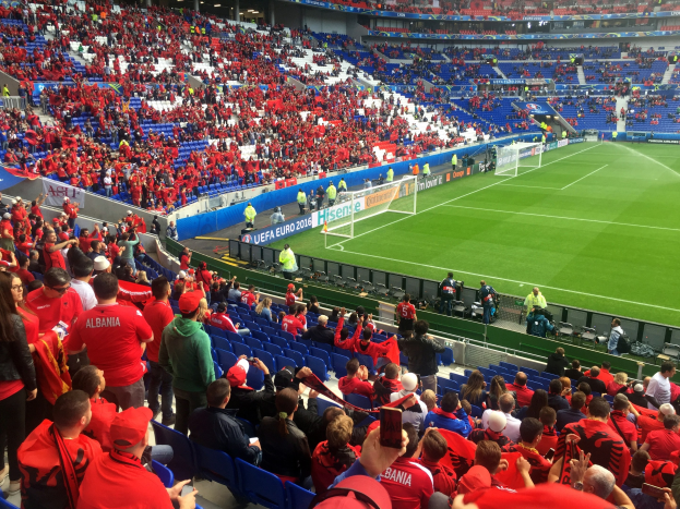 Eine große Menschenmenge in einem Stadion schaut ein Fußballspiel zu, mit Menschen auf Stühlen und Stehplätzen, Fahnen, einem Torpfosten und Deckenleuchten.