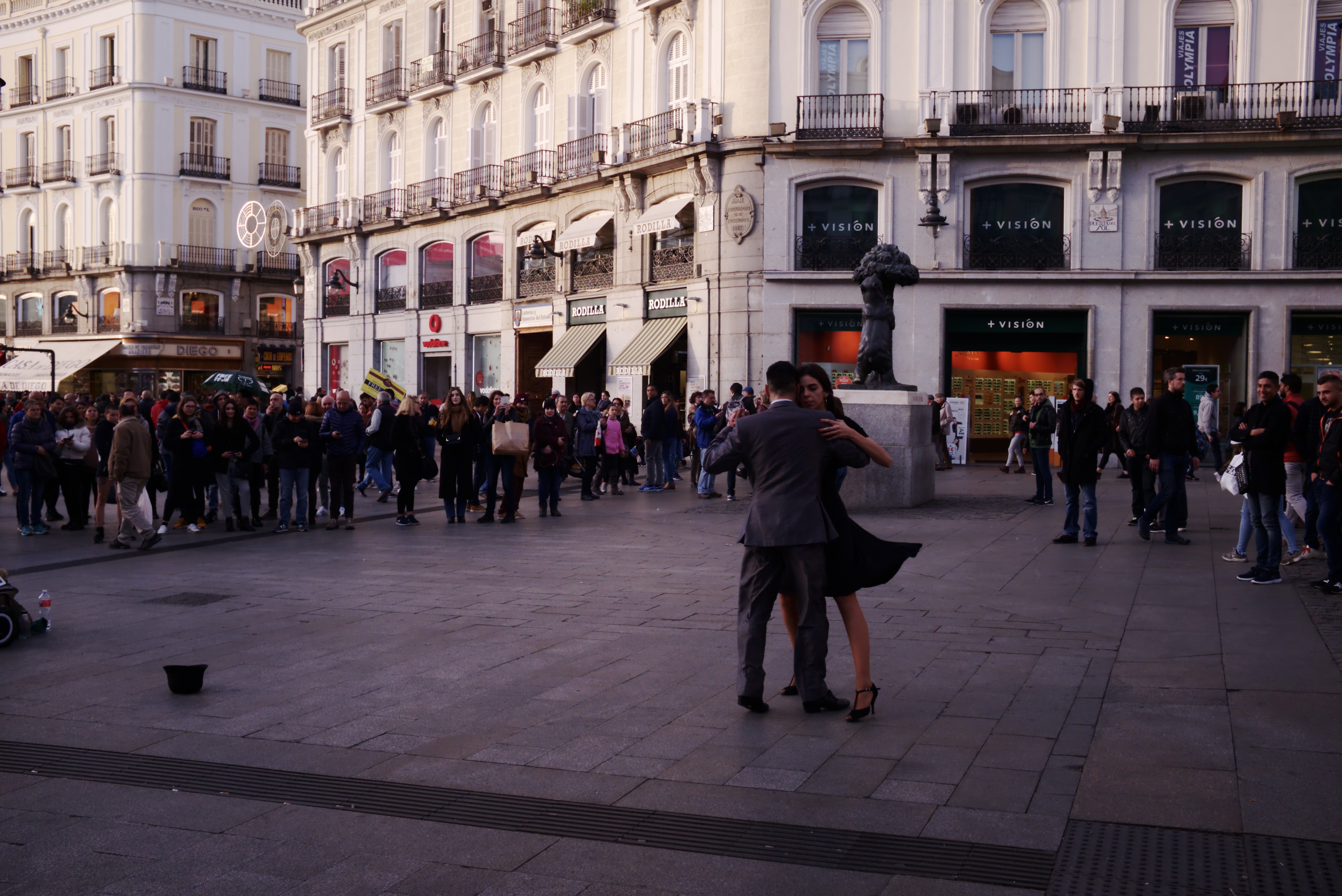 Ein Mann und eine Frau tanzen auf einem belebten Stadtplatz, mit einem Kinderwagen, Eimer und Polen links und Gebäuden, Geländern, Schildern, einer Statue und einem bewölkten Himmel im Hintergrund.