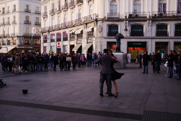Ein Mann und eine Frau tanzen auf einem belebten Stadtplatz, mit einem Kinderwagen, Eimer und Polen links und Gebäuden, Geländern, Schildern, einer Statue und einem bewölkten Himmel im Hintergrund.
