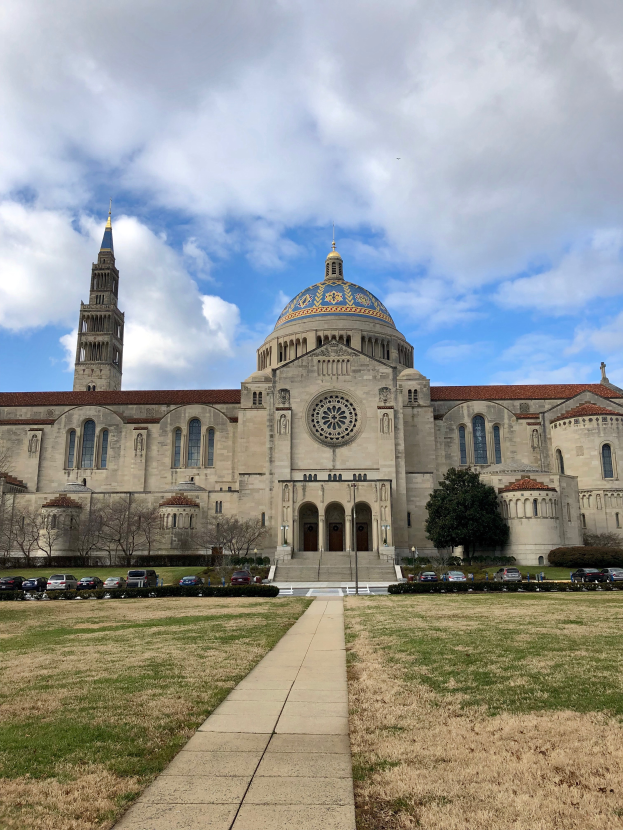 Die Basilika des Heiligen Herzens Jesu in St. Louis, Missouri, ein großes Gebäude mit Fenstern, Türen und Säulen, umgeben von einem Weg, Gras, Bäumen, Fahrzeugen, Pfosten und einem bewölkten Himmel.